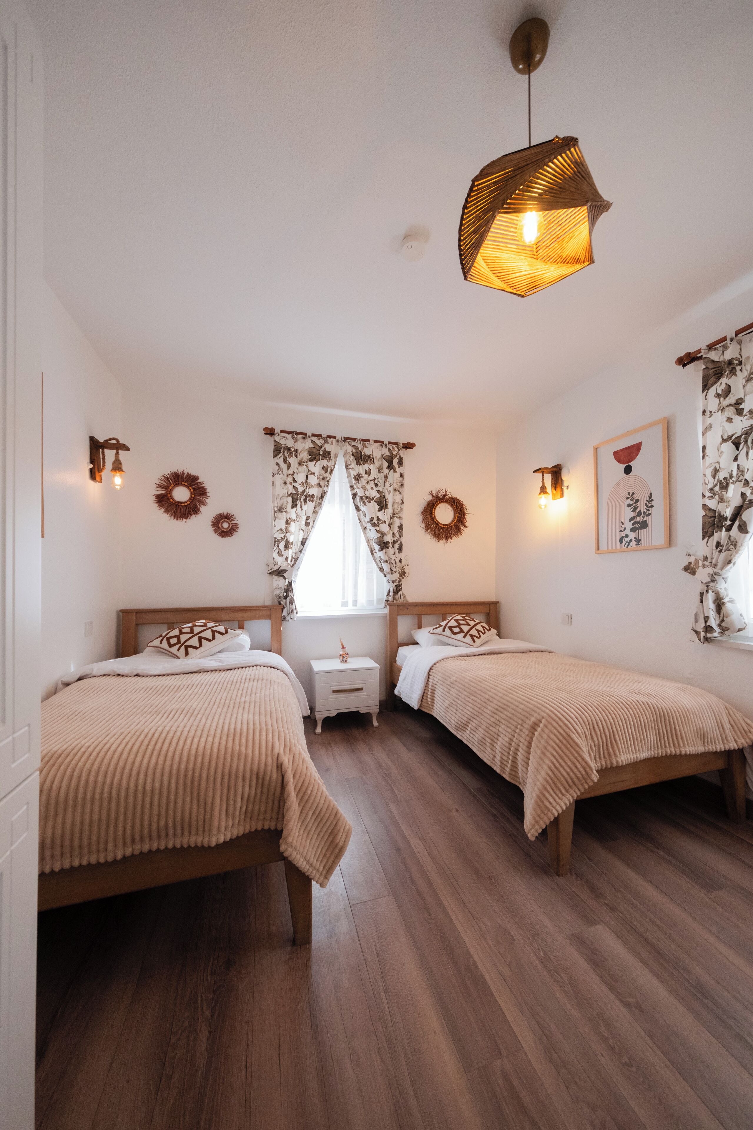 A bedroom with two beds, a nightstand, a hanging light fixture, and a window with floral curtains.