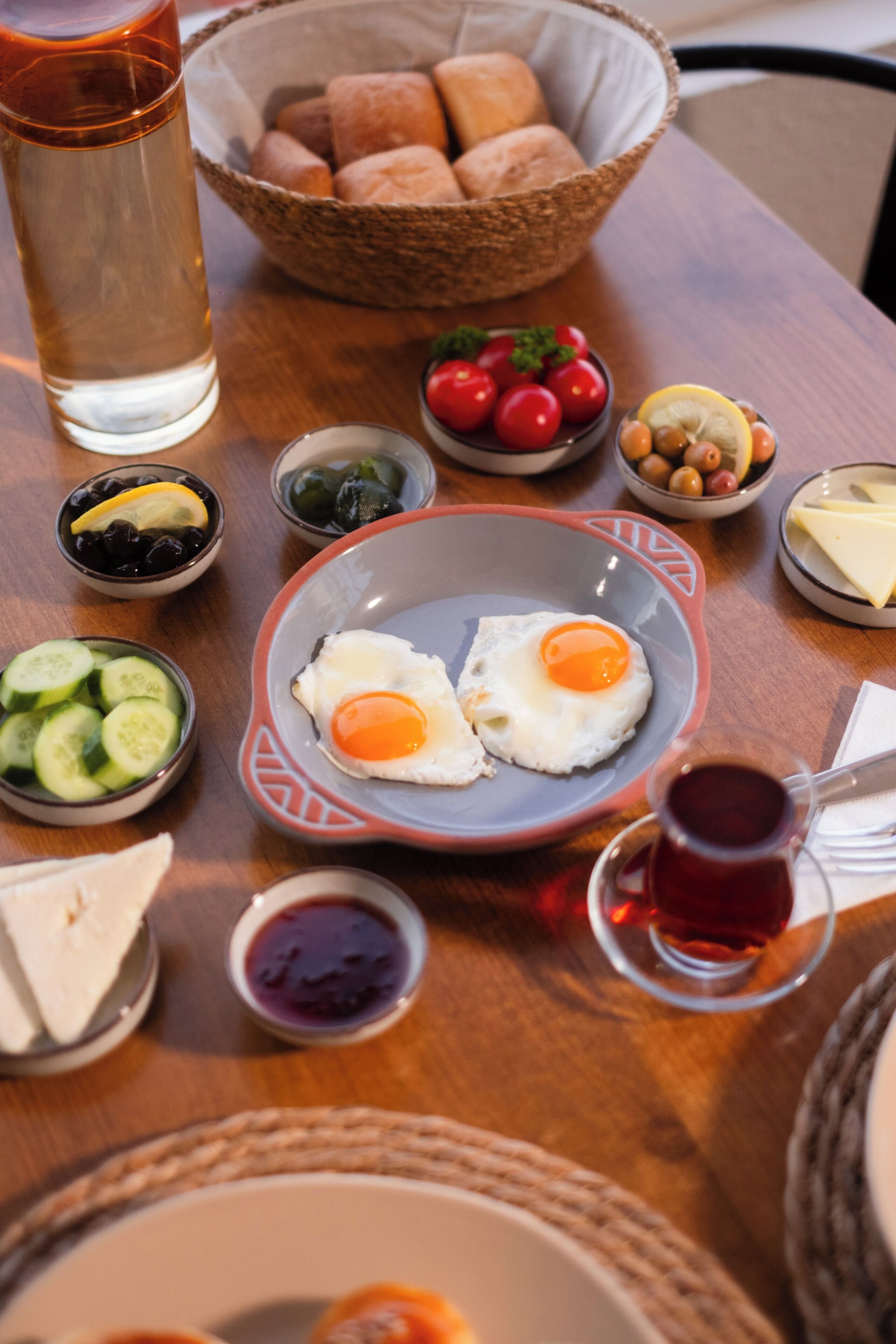 A breakfast table with eggs, bread, tomatoes, cucumbers, olives, and a glass of tea.