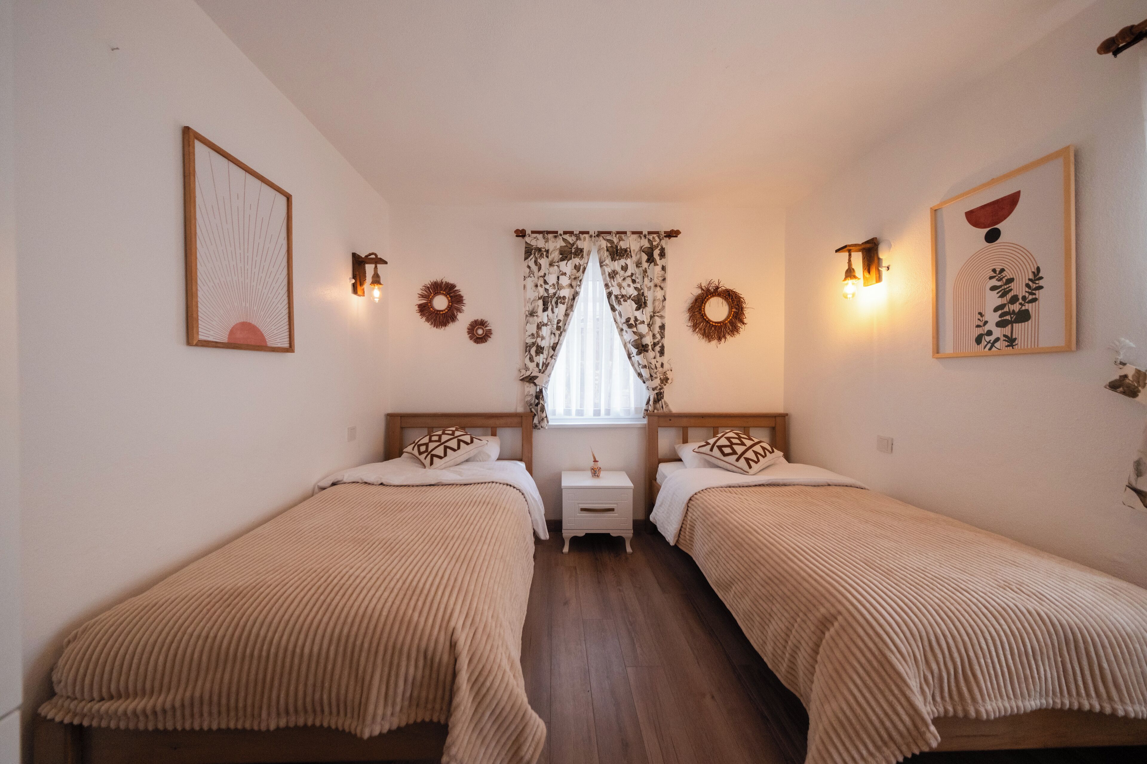 Una habitación con dos camas, una ventana con cortinas, luces fijadas a la pared y cuadros enmarcados.
