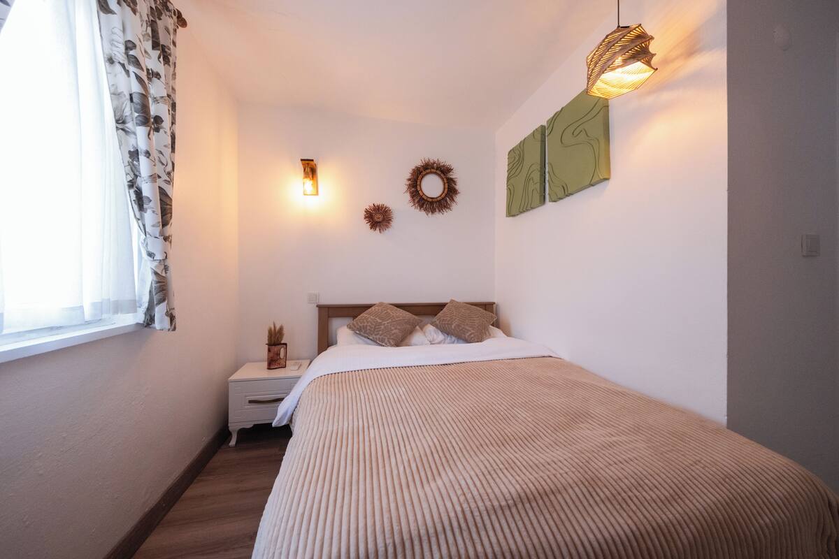 A bedroom with a bed, a nightstand, a wall-mounted lamp, and a window with curtains.