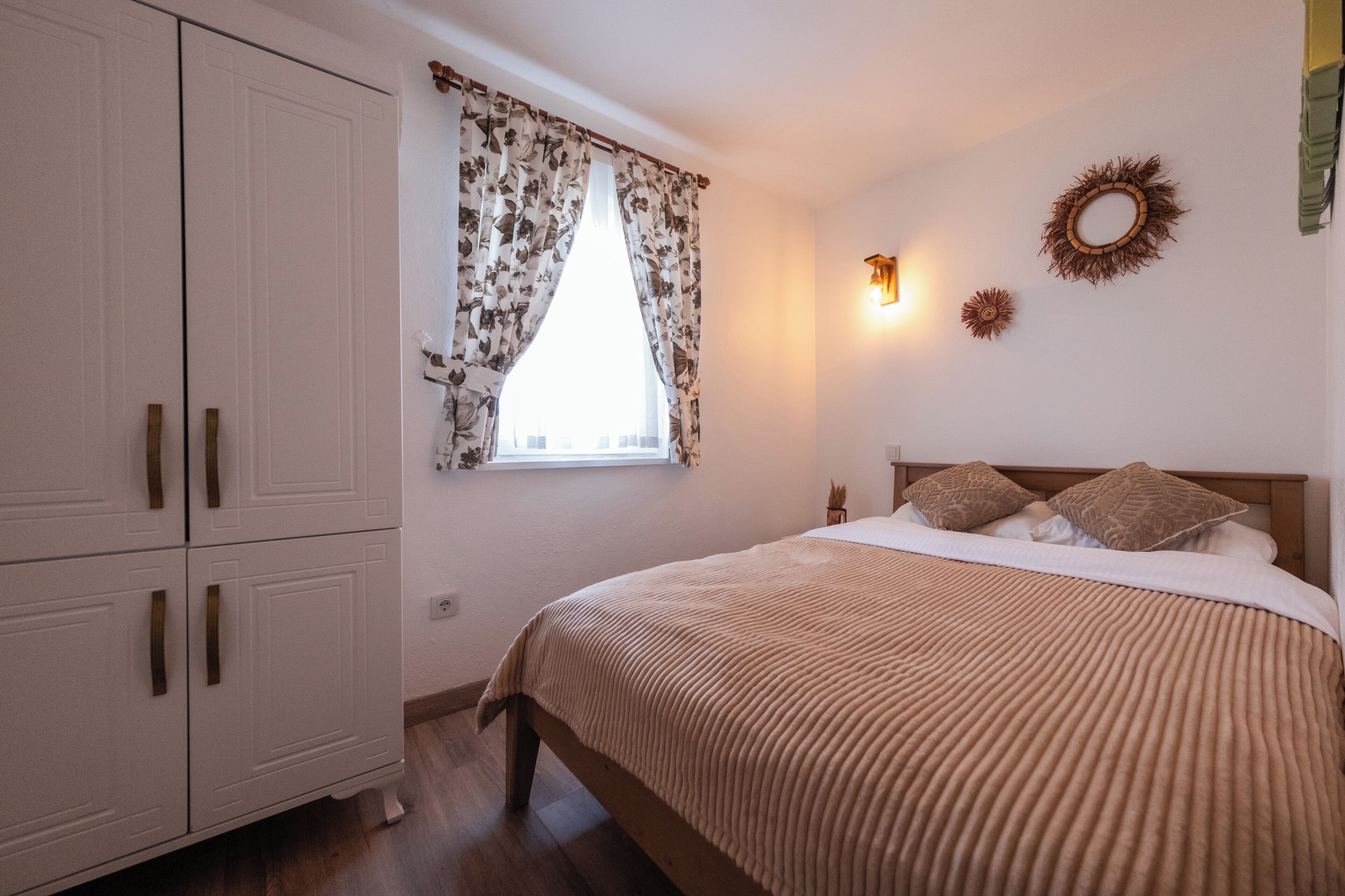 Ein Schlafzimmer mit einem Bett, einem Kleiderschrank, einem Fenster mit floralem Vorhang, Wandleuchten und einem dekorativen Kranz.