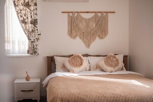 Un dormitorio con una cama, un colgador de macramé en la pared, una mesita de noche con un florero y una ventana con cortinas florales.