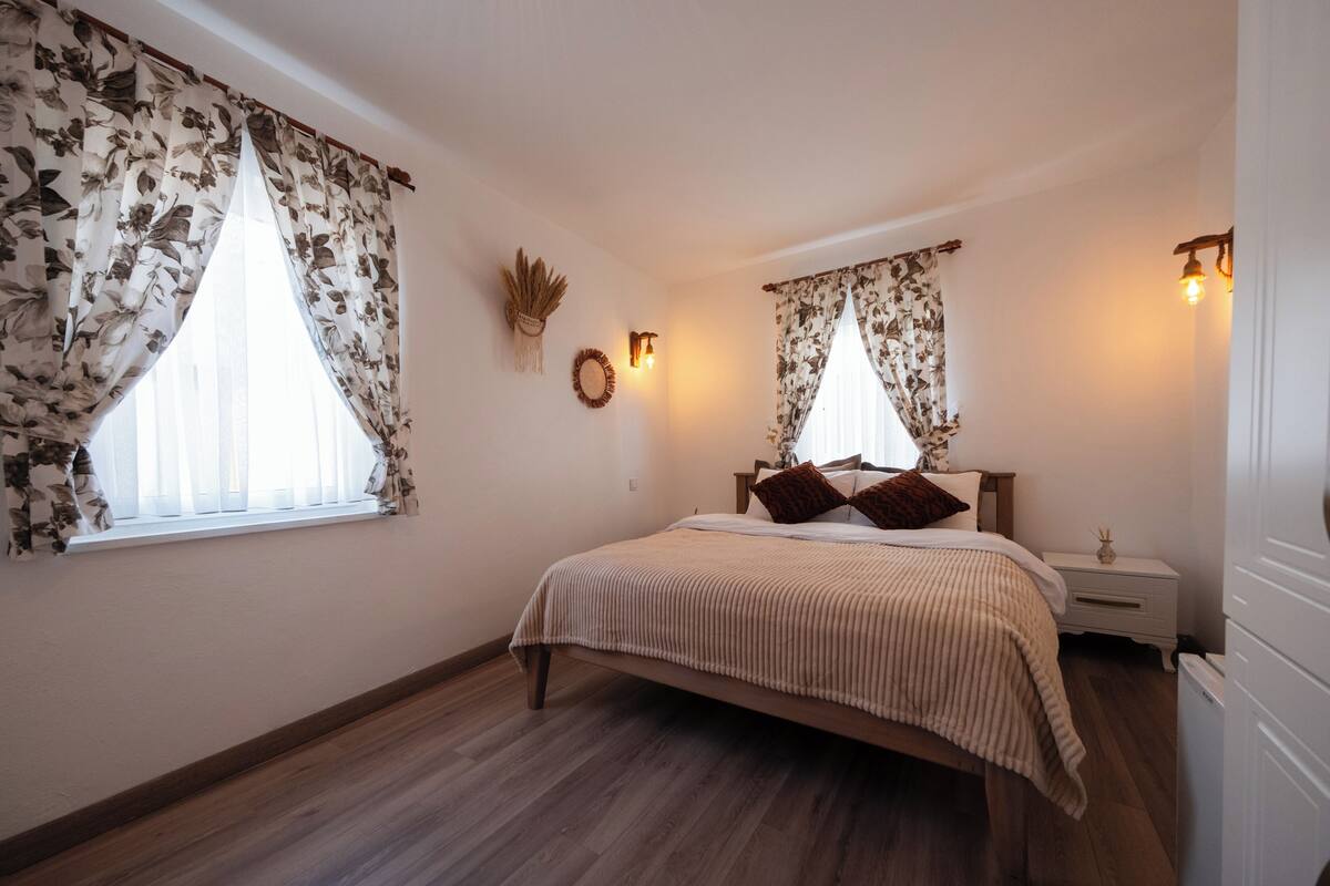 A bedroom with a bed, two nightstands, a wall-mounted light, and a window with floral curtains.