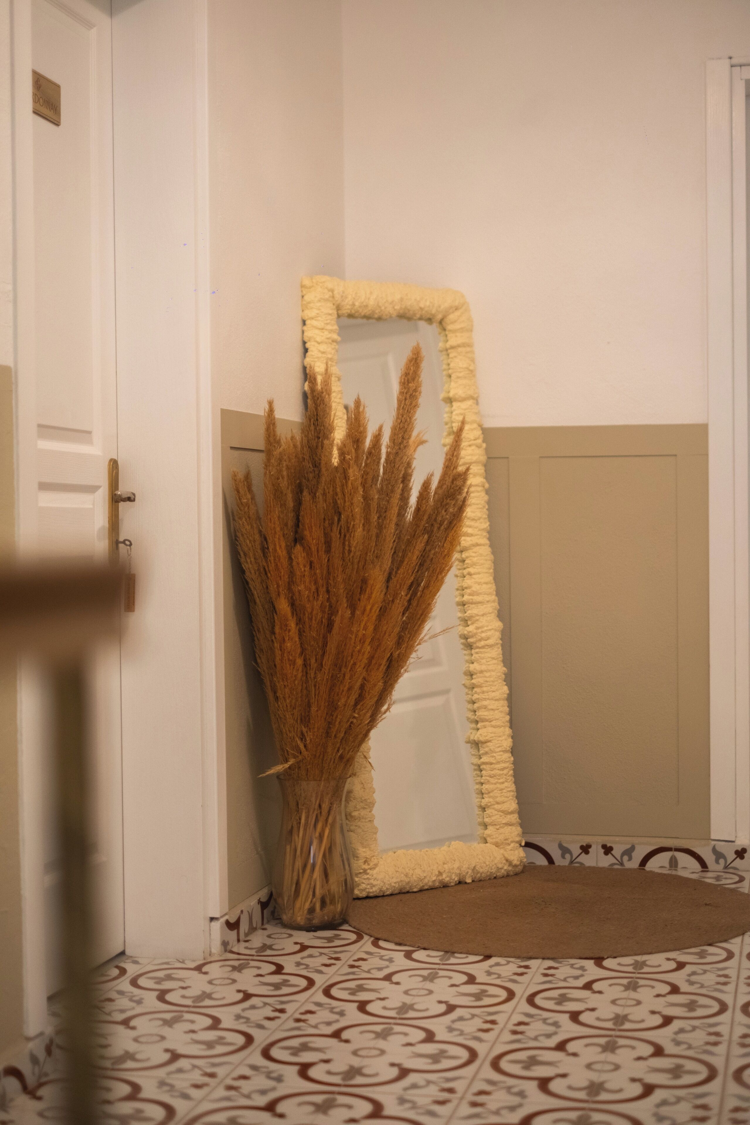 A mirror with a textured frame, a vase with dried plants, and patterned floor tiles.