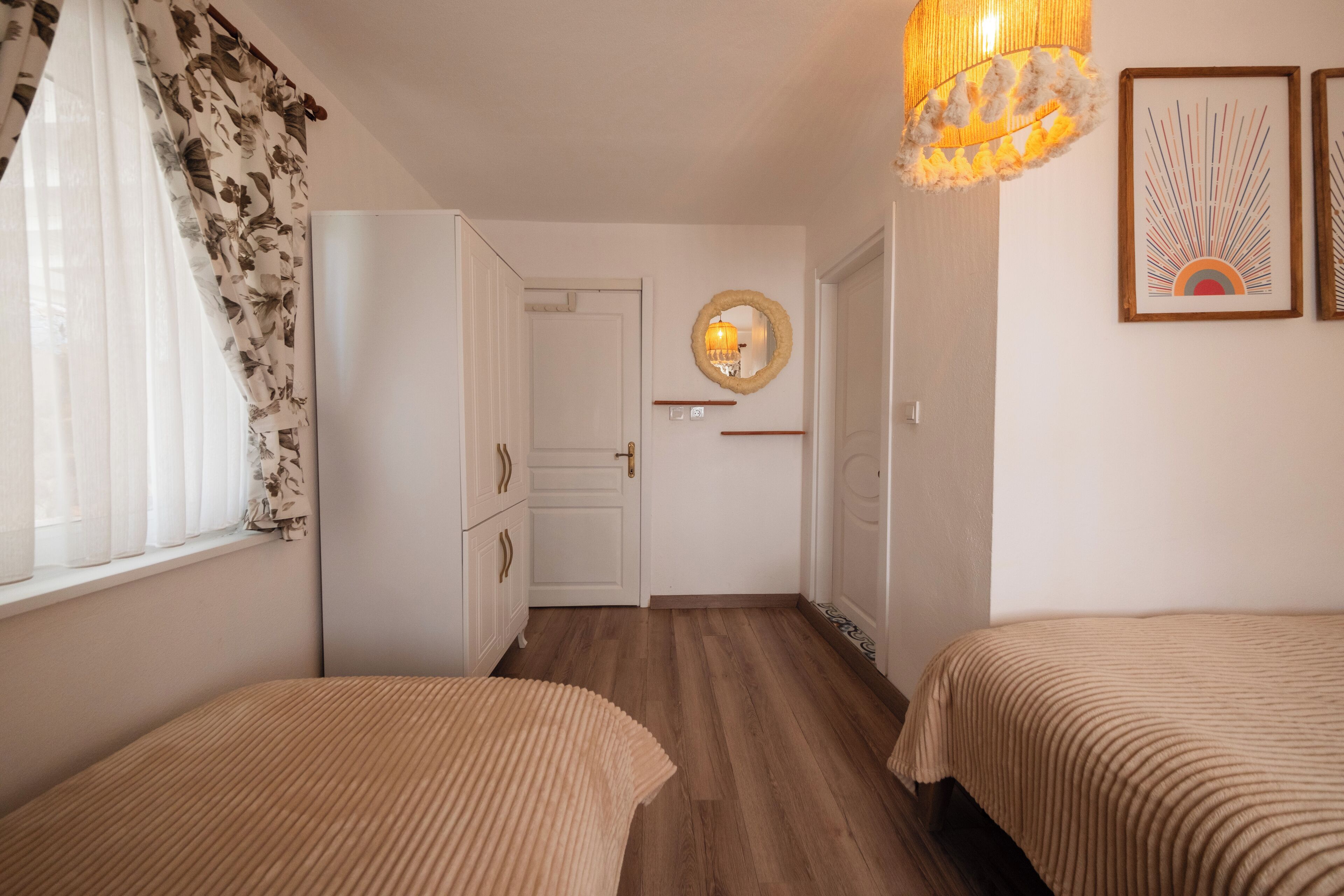 A hotel room with two beds, a wooden floor, a white wardrobe, a mirror, and a wall-mounted light fixture.