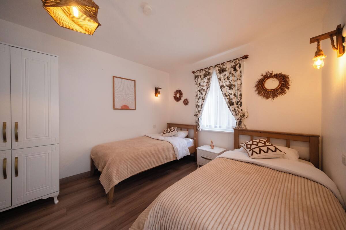 A bedroom with two single beds, a wardrobe, a nightstand, and a window with floral curtains.