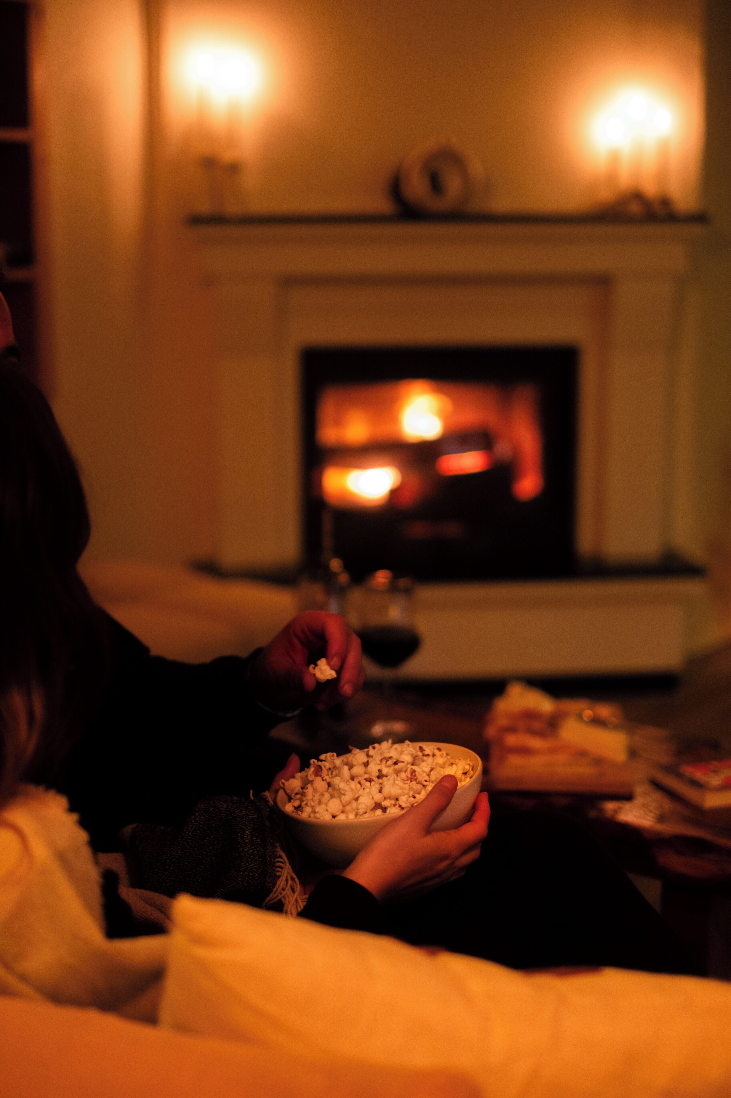 A person holding a bowl of popcorn and a glass of wine, sitting in front of a fireplace.