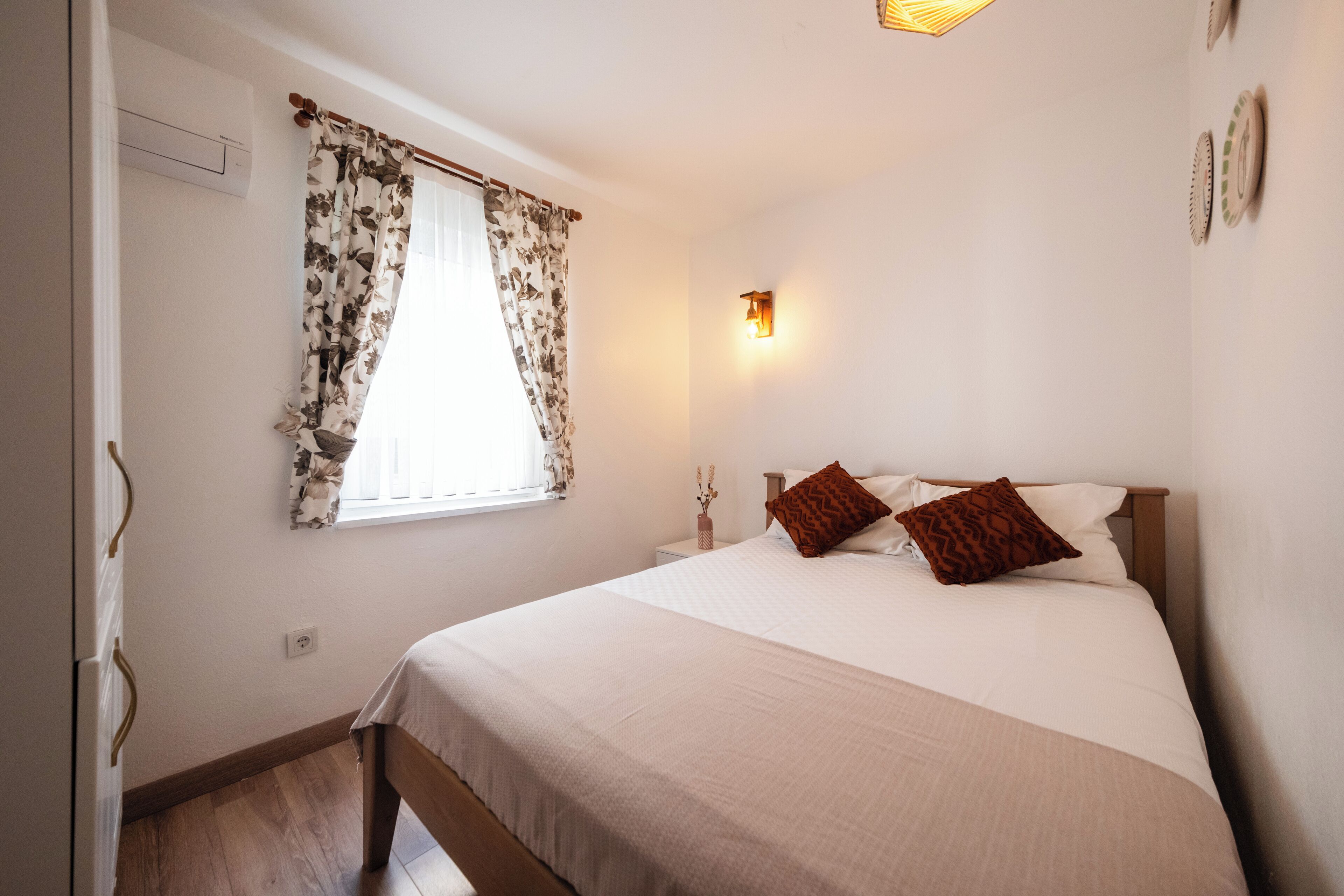 Ein Schlafzimmer mit einem Bett, einem Nachttisch, einer Wandleuchte und einem Fenster mit blumigen Vorhängen.