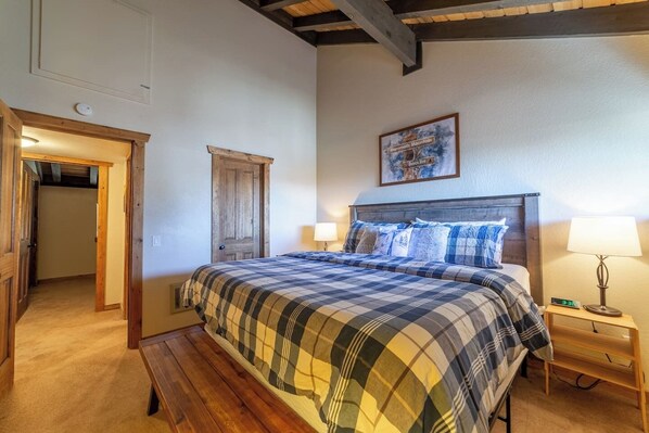 A bedroom with a bed, a bench, a nightstand, a wall clock, a door, and a framed picture.