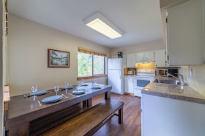 A dining area with a wooden table set for six, a kitchen with white cabinets and a built-in oven, and a window with a view of trees.