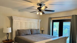 Une chambre avec une tête de lit blanche, un lit recouvert d’une couverture grise, deux lampes de chevet, une table de chevet avec des tiroirs et un ventilateur de plafond.
