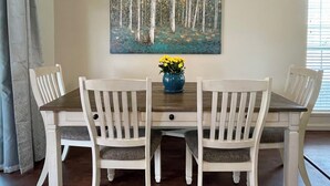 Une table à manger entourée de quatre chaises blanches, un vase bleu contenant des fleurs jaunes et un tableau représentant une forêt accroché au mur.