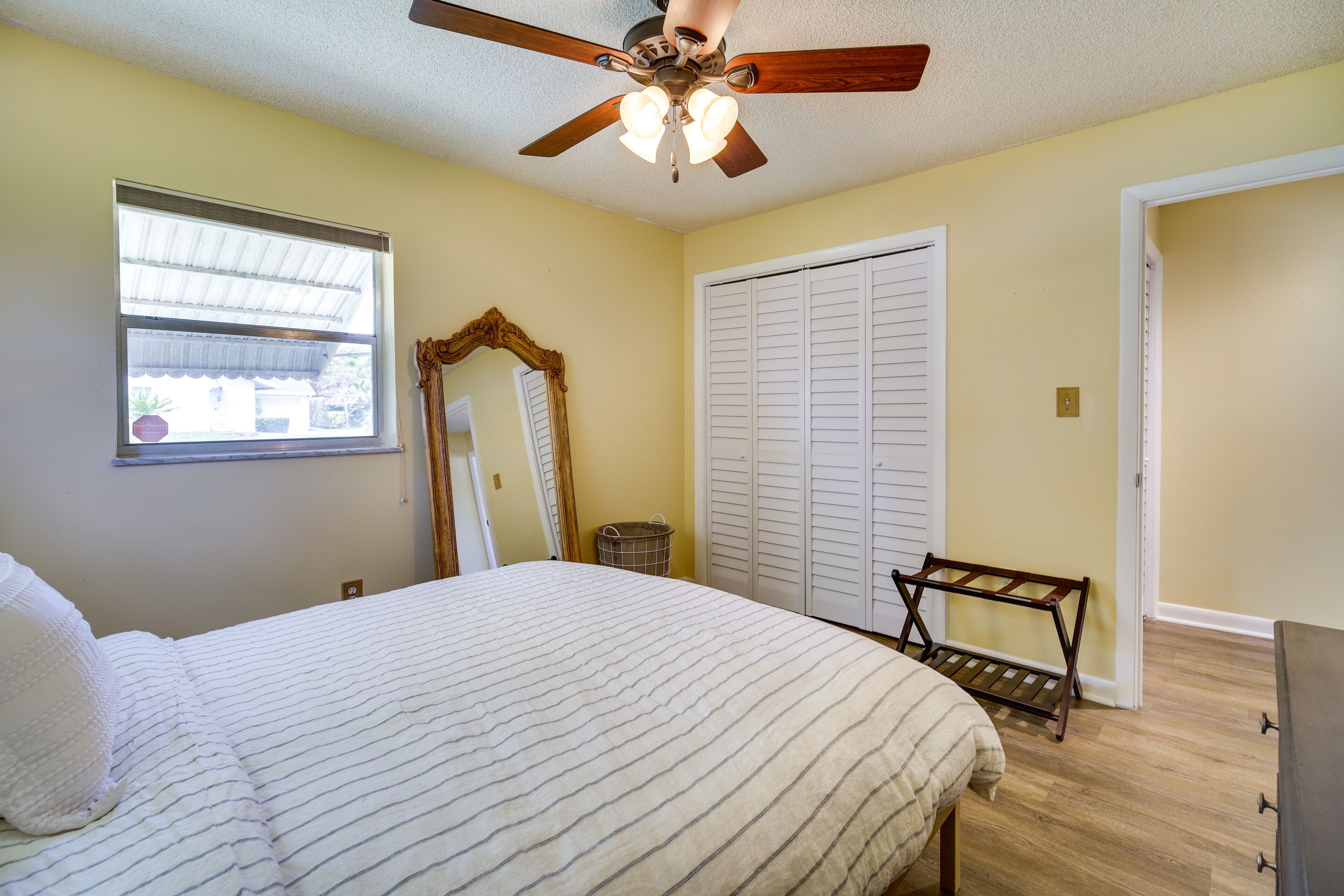 Une chambre à coucher comprenant un lit, un ventilateur de plafond, une fenêtre, un miroir, une armoire et un banc.