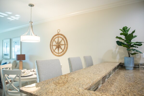 Un comedor con una mesa larga, sillas, una lámpara colgante, una brújula en la pared y una planta en maceta.