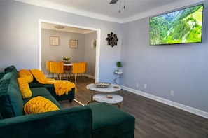 Sala de estar com sofá verde, televisão fixada na parede e área de jantar com cadeiras amarelas.
