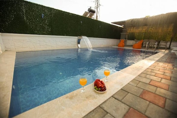 Une piscine agrémentée d’une cascade, deux verres de jus d’orange et un saladier de fruits sur le bord.