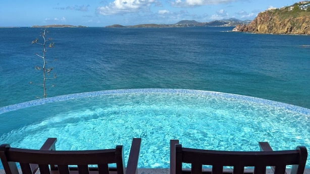 An infinity pool with two wooden chairs facing the ocean, under a clear sky.