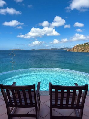 Piscina infinita com duas cadeiras de madeira voltadas para o oceano, sob um céu claro.