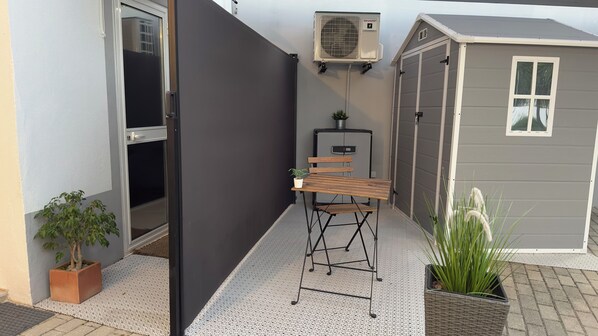 Ein kleiner Außenbereich mit einem Metallstuhl, einem Blumentopf, einem grauen Geräteschuppen und einer an der Wand befestigten Klimaanlage.