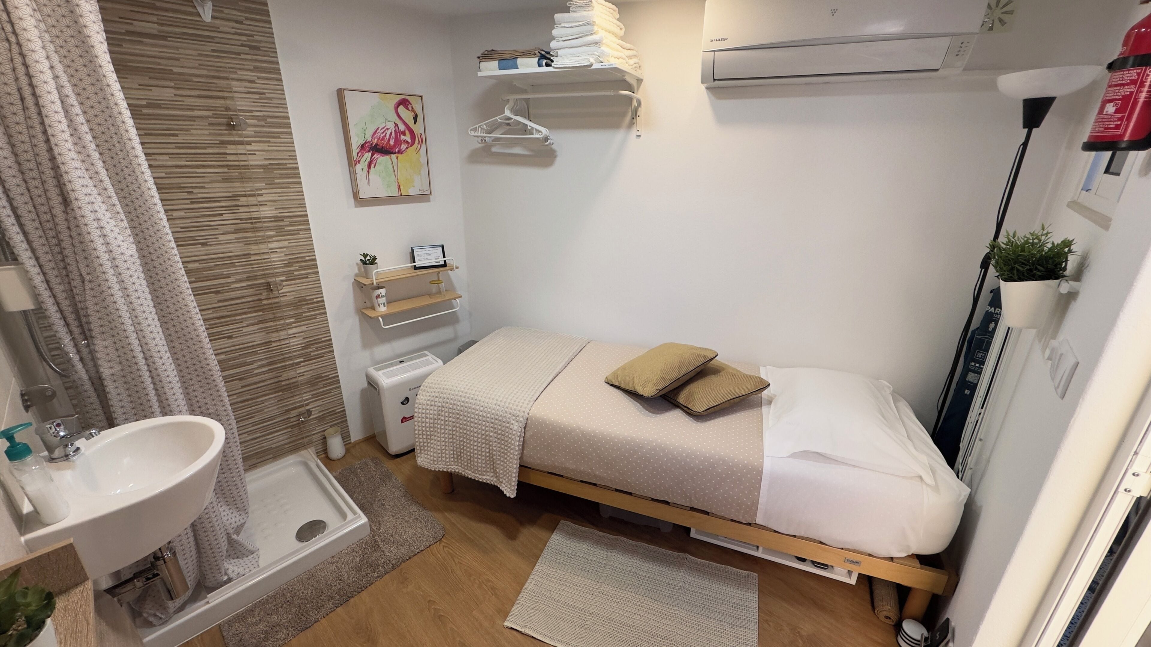 A compact bedroom with a bed, a bedside table, a wall-mounted shelf, a small bedside table, a radiator, a window with curtains, a bathroom with a sink, and a shower area.