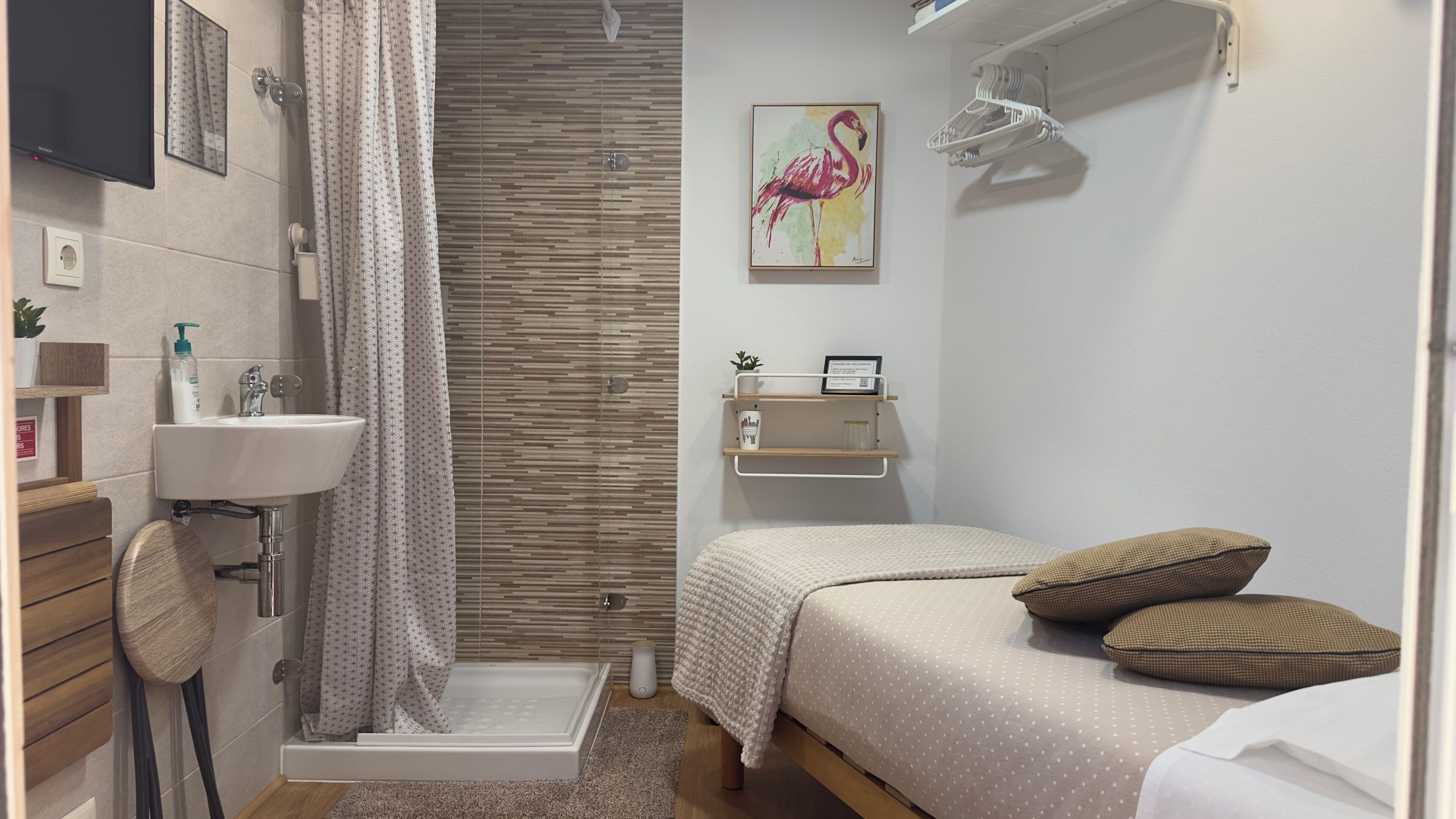 A small, modern bedroom with a shower, a sink, a bed with pillows, a shelf with toiletries, and a painting of a flamingo.