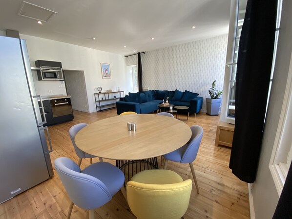 A modern kitchen with a refrigerator, microwave, and stove. There is a dining area with a round table and chairs. A blue sofa and a small side table are in the living area.