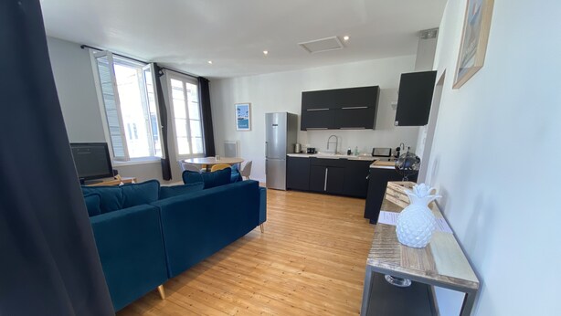 Una sala moderna con un sofá azul, piso de madera y una cocina con armarios negros y encimera blanca.