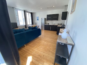A modern living room with a blue sofa, wooden flooring, and a kitchen area with black cabinets and a white countertop.
