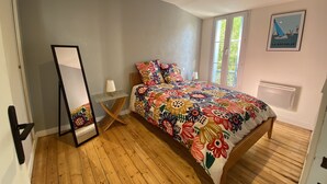 Un dormitorio con piso de madera, una cama con colcha floral de colores, una mesita de noche con lámpara, un espejo y una ventana con vista a vegetación.