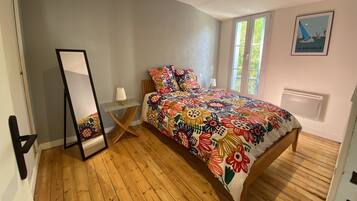 Une chambre avec un plancher en bois, un lit recouvert d’une courtepointe à motifs floraux colorés, une table de chevet avec une lampe, un miroir et une fenêtre donnant sur un paysage verdoyant.