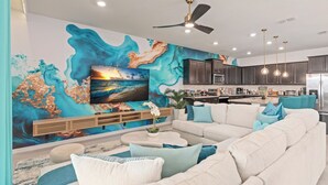 A modern living room with a large wall mural, a flat-screen TV, a ceiling fan, and a kitchen area in the background.