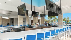 A bar area with white bar counters, blue cushioned chairs, and multiple flat-screen TVs mounted on the ceiling.