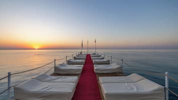 A wooden pier with white beds lined up, a red carpet, and a view of the sunset over the water.
