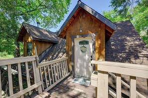A wooden cabin with a white door and a welcome mat, surrounded by a wooden deck and trees. - 'Lakeside Bear Lodge' Studio: 9 Mi to Bass Lake! (Wishon)
