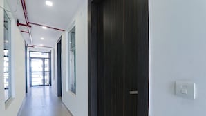 A long, narrow hallway with a dark wooden door on the right, a light switch, and a window on the left. - Fabhotel The Comfort (Prayagraj)