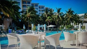 A poolside area with lounge chairs and umbrellas, palm trees, and a hotel building in the background.
