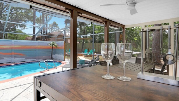 A poolside area with a wooden deck, a glass table with two wine glasses, and a view of a swimming pool and outdoor furniture. - Unique A-Frame Heated pool & Firepit  (Cocoa)