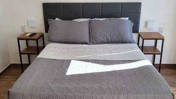 Ein ordentlich bezogenes Bett mit einer grauen Tagesdecke, zwei Nachttische mit Lampen, ein dunkles Kopfteil und ein an der Wand hängendes abstraktes Kunstwerk.