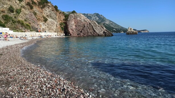 A pebble beach with clear water, rocky cliffs, and a distant mountain. - ACIKA apartments Rafailovici (Becici)