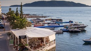 A waterfront restaurant with outdoor seating, boats docked nearby, and a view of the sea and distant hills. - ACIKA apartments Rafailovici (Becici)