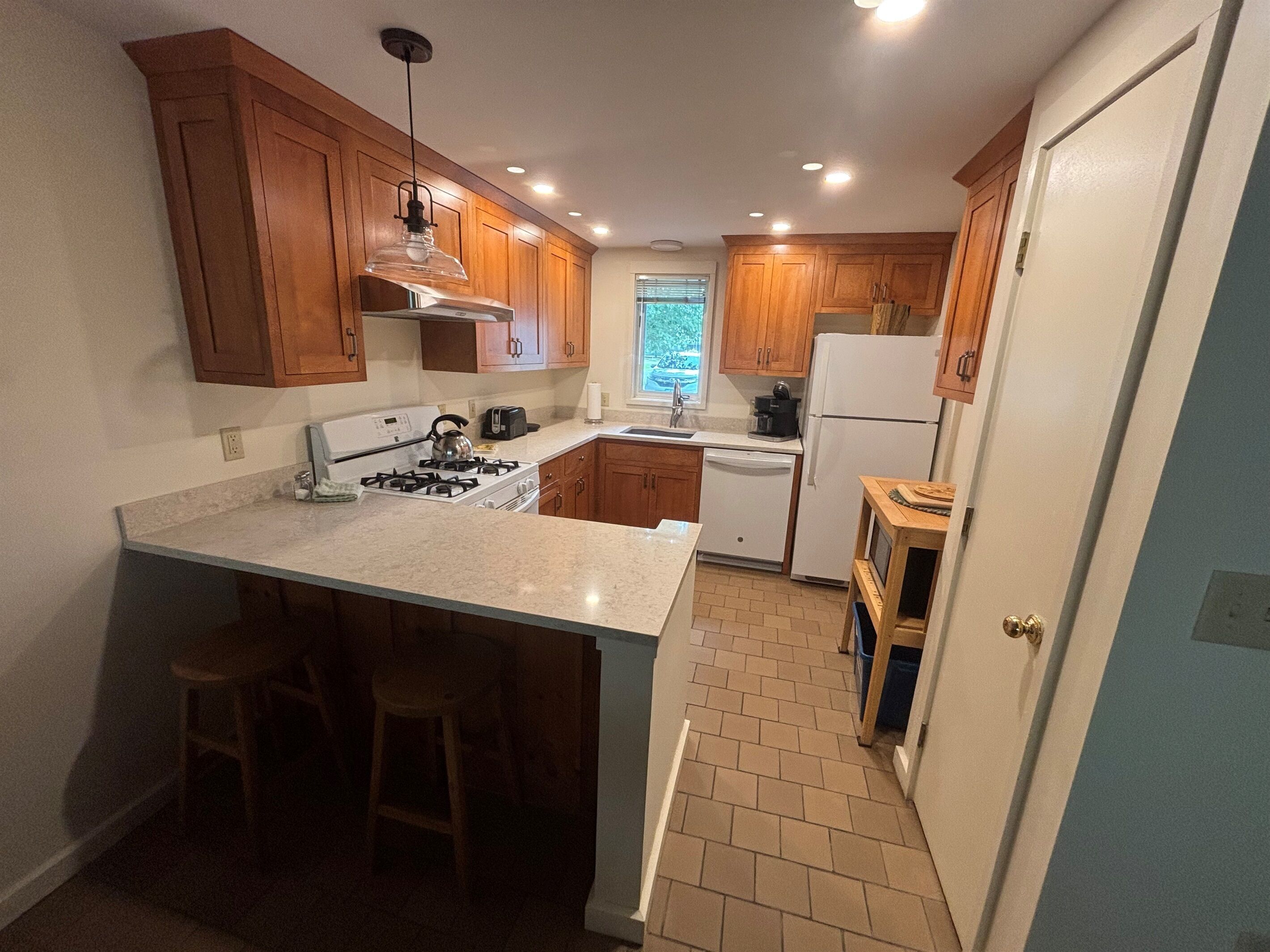 A compact kitchen with wooden cabinets, a central island with stools, a stove, a refrigerator, and a window providing natural light.