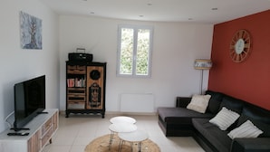 Una sala de estar con televisor, un mueble de madera con libros, un reloj de pared rojo y una ventana con luz natural.
