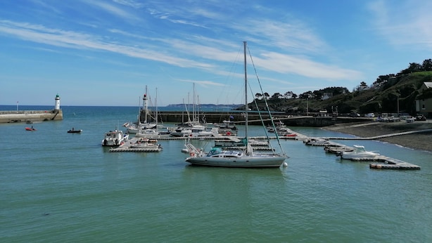 Puerto deportivo con barcos amarrados, un faro y un paisaje montañoso al fondo.