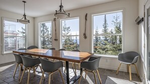Un espace repas avec une table et des chaises en bois, de grandes fenêtres donnant sur la verdure et des luminaires suspendus.
