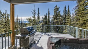 Un espace extérieur abrité, doté d’un barbecue et d’un bain à remous, entouré d’une rambarde en métal et offrant une vue sur une zone montagneuse boisée.