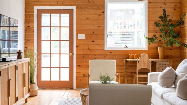 Una habitación acogedora con paredes de madera, un ventanal grande, un sofá, una silla y un televisor sobre un mueble.