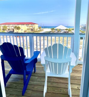 Relax in Adirondack chairs on a deck overlooking a sandy beach and palm trees.