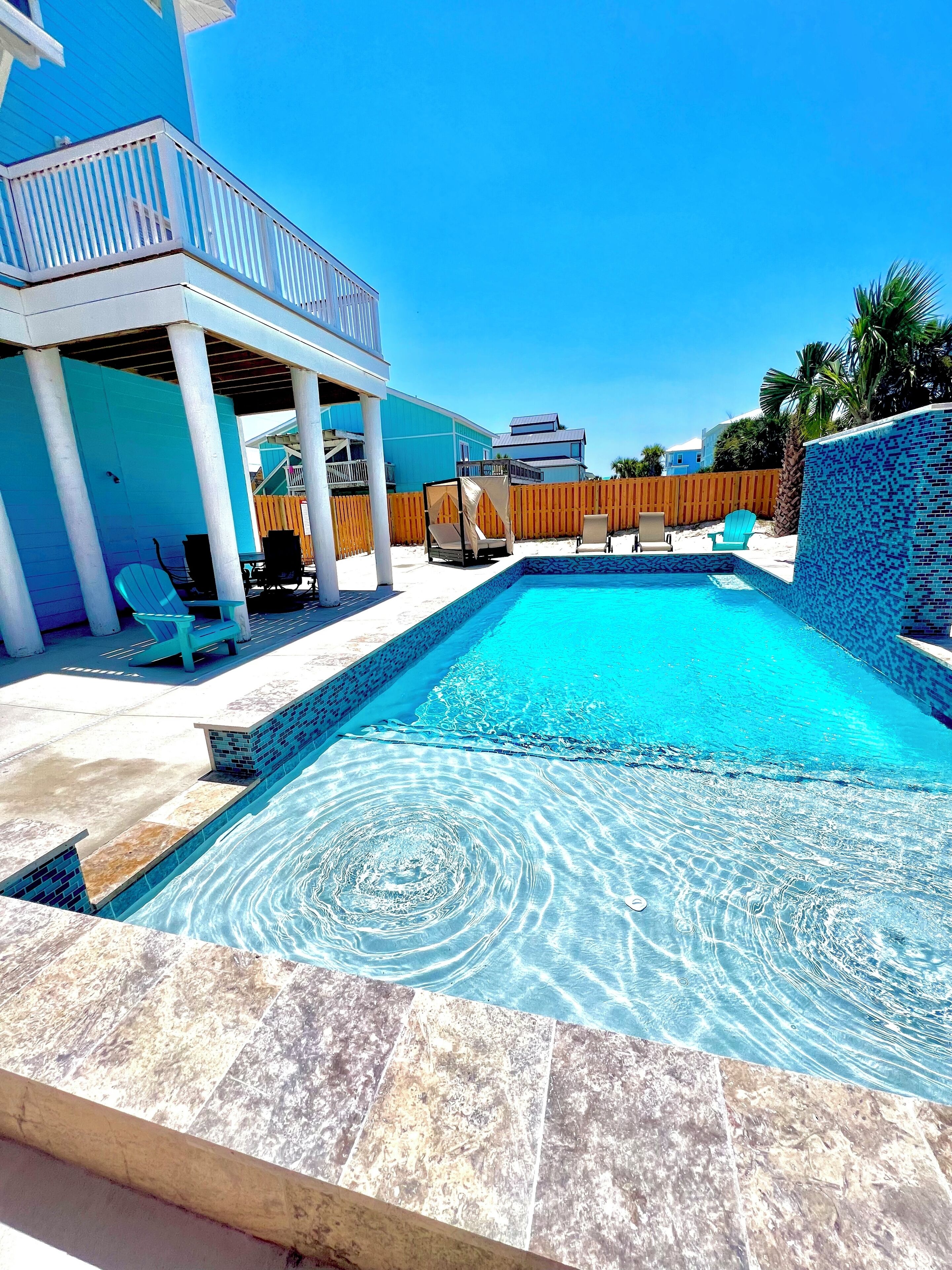 A swimming pool with a tiled edge, surrounded by a covered patio area with chairs and a wooden fence.