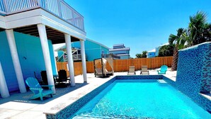 A swimming pool with a tiled edge, surrounded by a covered patio area with chairs and a wooden fence. - Luxury Gulf-View Beach Retreat Pool, Deck, Steps to Sand, Pet-Friendly, Sleeps 9 (Gulf Breeze)