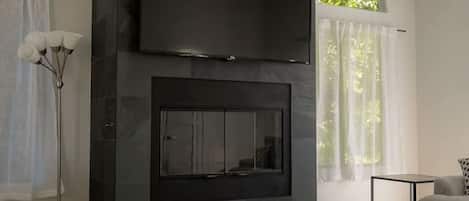 Uma sala de estar moderna com uma TV de tela plana fixada em uma parede escura, uma mesa de centro de vidro, um tapete estampado e um sofá.